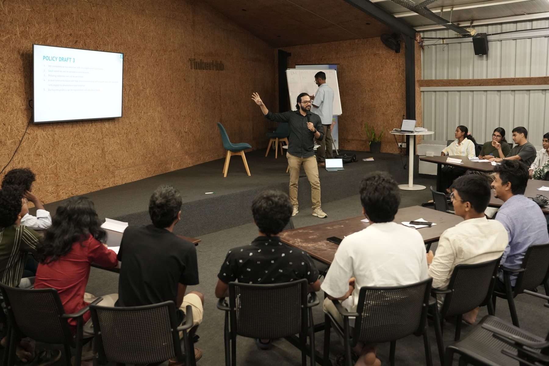 An NPoC co-founder leads the policy simulation game, with a policy draft on screen and participants seated in stakeholder groups